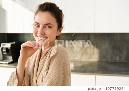 Portrait of beautiful and happy woman standing in kitchen in bathrobe, posing and smiling at camera, cooking food, preparing meal, eating lunch near counter Portrait of beautiful and happy woman standing in kitchen in bathrobe, posing and smiling at camera, cooking food, preparing meal, eating lunch near counter 107739244