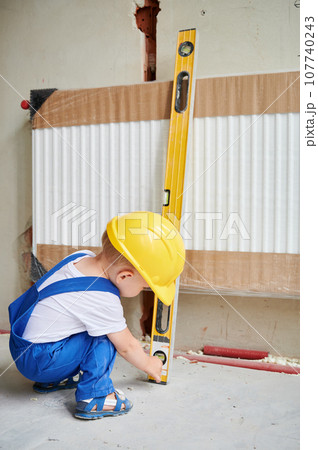 Child in safety helmet using spirit level ruler while installing heating radiator in apartment under renovation. Kid construction worker checking precision measuring device at home. 107740243