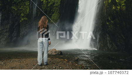 Tourist girl raising arms standing at powerful waterfall Air Terjun Munduk, Indonesia, Bali island. Woman posing, back view. Beautiful nature landscape. Travel, tourism, holiday Tourist girl raising arms standing at powerful waterfall Air Terjun Munduk, Indonesia, Bali island. Woman posing, back view. Beautiful nature landscape. Travel, tourism, holiday 107740372