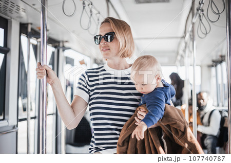 Mother carries her child while standing and holding on to bar holder on bus. Mom holding infant baby boy in her arms while riding in public transportation. Cute toddler traveling with mother 107740787