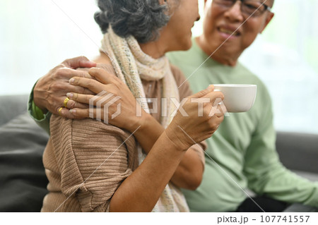 Carefree middle aged old husband embracing wife, mature woman holding cup of tea, relaxing on sofa Carefree middle aged old husband embracing wife, mature woman holding cup of tea, relaxing on sofa 107741557