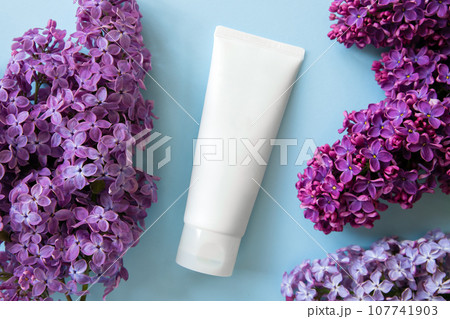 Cosmetic cream moisturizer in tube bottle and purple lilac flowers a lot on blue background. Unbranded lotion, balsam, hand creme, toothpaste, container of cream, skin care. Packaging mockup close-up 107741903