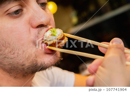 Young adult male man eating baked Japanese sushi roll with bamboo chopsticks at sushi restaurant. Tempura fried roll and wasabi sauce. Male unshaven face with roll in his mouth close-up. Seafood. 107741904