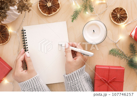 Christmas wish list writing. Woman creating present list for winter holidays. Top view, flat lay. Preparing for Xmas and New Year concept. Female hand writing in empty notebook on wooden table 107746257