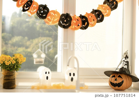 Halloween concept. A pumpkin with a painted face, a white ghost and a bouquet of yellow chrysanthemum flowers in a black vase against the background of a window in home interior. 107751814