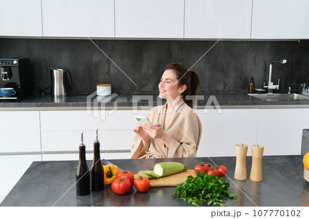 Portrait of woman in bathrobe sitting in kitchen with smartphone, cooking dinner, watching recipe on social media, video tutorial how to prepare meal Portrait of woman in bathrobe sitting in kitchen with smartphone, cooking dinner, watching recipe on social media, video tutorial how to prepare meal 107770102