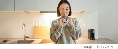 Portrait of smiling asian woman standing on her kitchen, drinking coffee and looking at camera, concept of cozy and comfortable home 107770141