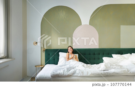 Vertical shot of beautiful asian girl lying in bedroom with sophisticated interrior, smiling and looking happy in morning, spending time in bed 107770144