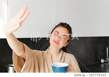 Portrait of beautiful, happy young woman covers her eyes from morning sunlight, waking up and drinking coffee, holding mug, posing in the kitchen Portrait of beautiful, happy young woman covers her eyes from morning sunlight, waking up and drinking coffee, holding mug, posing in the kitchen 107770145