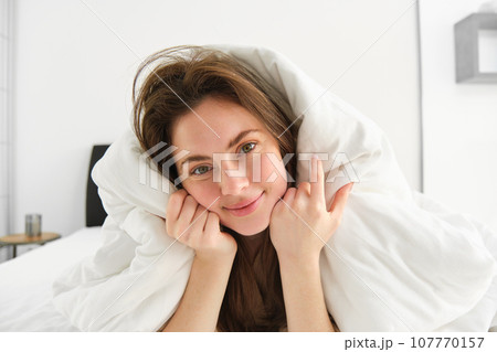 Portrait of gorgeous smiling woman, lying in bed covered in duvet, has messy hair, looking happy, relaxing in her bedroom, spending time in hotel room in morning 107770157