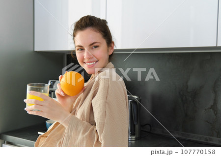Portrait of happy woman in kitchen, wearing bathrobe, drinking orange juice, freshly squeezed drink, smiling and laughing, food and drink concept 107770158