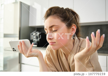 Attractive young cheerful girl baking at the kitchen, making dough, holding recipe book, having ideas 107770159