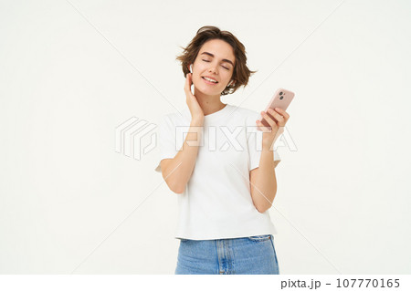 Portrait of smiling, beautiful young woman in wireless headphones, puts on music in earphones, holds smartphone, using streaming app, standing over white background 107770165