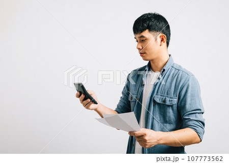 A young man, appearing sad and tired, strains while holding a paper and calculator, meticulously calculating bills. He is visibly worried over the financial matter. Isolated on a white background. 107773562