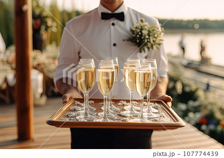 Waiter at a party, carrying a tray with champagne or wine glasses. Outdoor wedding Waiter at a party, carrying a tray with champagne or wine glasses. Outdoor wedding 107774439
