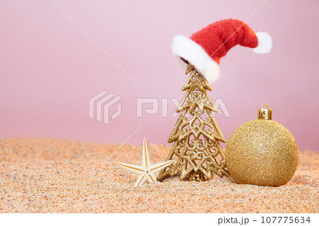 Golden Christmas tree in Santa's hat with shiny ball and starfish on sand of beach. New Year in hot countries. Copy space Golden Christmas tree in Santa's hat with shiny ball and starfish on sand of beach. New Year in hot countries. Copy space 107775634