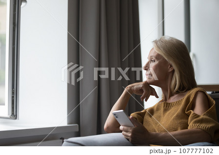 Thoughtful serious older woman holding smartphone, thinking over bad news 107777102