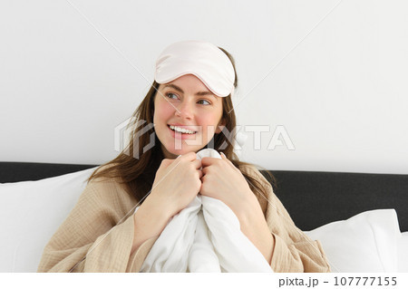 Pretty girl wakes up in morning, lying on pillow covered with white duvet, smiling at camera, wakes-up and looks happy 107777155