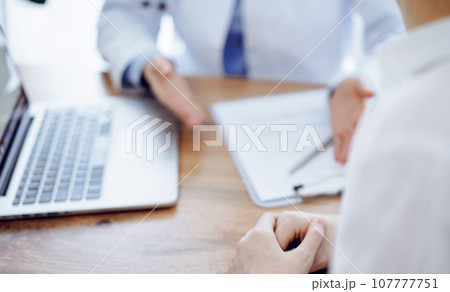 Doctor and patient sitting and discussing something at wooden table while using a laptop computer. Focus is on patient's hands. Medicine concept 107777751