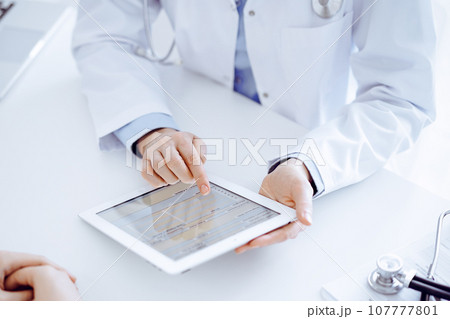 Doctor and patient sitting opposite each other at the desk in clinic. The focus is on female physician's hands pointing into tablet computer touchpad, close up. Medicine concept 107777801