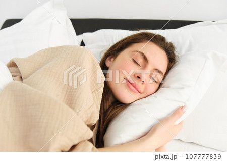 Portrait of smiling brunette woman in pyjamas, sleeping in hotel bed, relaxing with pleased face, dreaming, sleeping in bedroom 107777839