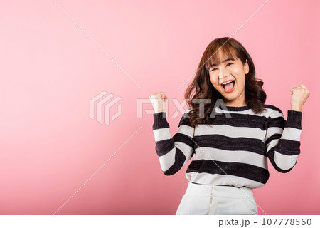 Portrait of a jubilant Asian woman with raised fists, celebrating her success with a happy expression. Studio shot isolated on a pink background, conveying victory and joy. Portrait of a jubilant Asian woman with raised fists, celebrating her success with a happy expression. Studio shot isolated on a pink background, conveying victory and joy. 107778560