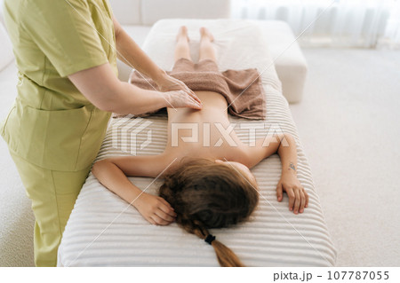Closeup top view of unrecognizable female pediatric masseuse massaging back of ten-year-old girl lying on couch in medical clinic. Little child having therapeutic, preventive massage session. Closeup top view of unrecognizable female pediatric masseuse massaging back of ten-year-old girl lying on couch in medical clinic. Little child having therapeutic, preventive massage session. 107787055