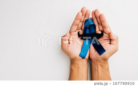 November highlights men's health and Prostate cancer awareness. A man's hands grip a light blue ribbon with a mustache on a blue background a symbol of strength. 107790910