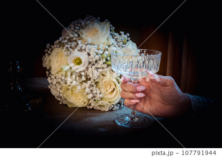 White rose bridal bouquet on the table. The bride is holding a crystal glass White rose bridal bouquet on the table. The bride is holding a crystal glass 107791944