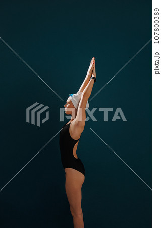 Side view of a young slim female swimmer preparing for competition standing against a blue backdrop 107800389