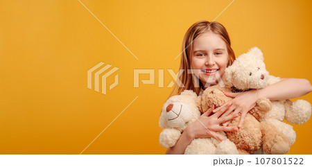 Beautiful girl child with three teddy bears in her hands isolated on yellow background with copyspace. Horizontal portrait of kid holding toys and smiling looking at the camera 107801522