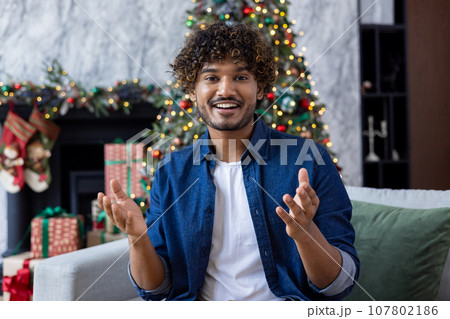 Online video call, man recording holiday greetings to friends and relatives, hispanic looking at webcam smiling and talking, sitting on sofa at Christmas, celebrating new year, with tree background. 107802186