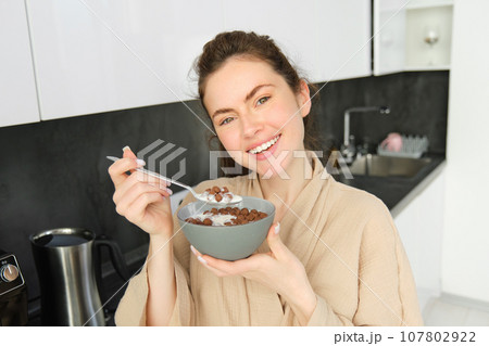 Close up of beautiful smiling woman in bathrobe, standing in kitchen near worktop, eating bowl of chocolate cereals with milk, holding spoon and looking happy at camera Close up of beautiful smiling woman in bathrobe, standing in kitchen near worktop, eating bowl of chocolate cereals with milk, holding spoon and looking happy at camera 107802922