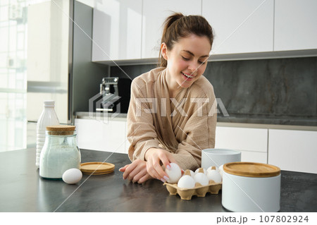Attractive young cheerful girl baking at the kitchen, making dough, holding recipe book, having ideas 107802924