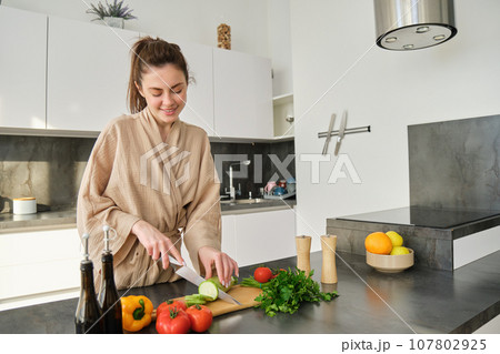 Portrait of good-looking woman cooking salad in the kitchen, chopping vegetables and smiling, preparing healthy meal, leading healthy lifestyle and eating raw food Portrait of good-looking woman cooking salad in the kitchen, chopping vegetables and smiling, preparing healthy meal, leading healthy lifestyle and eating raw food 107802925