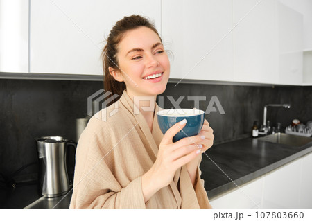Portrait of happy young woman starts her morning with mug of coffee, drinking tea from cup, standing in the kitchen, smiling cheerfully 107803660