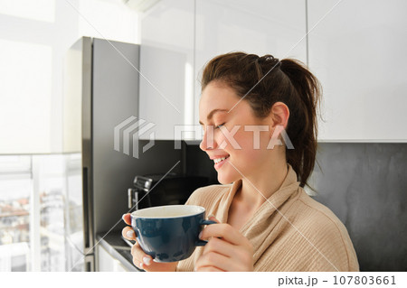 Portrait of happy young woman starts her morning with mug of coffee, drinking tea from cup, standing in the kitchen, smiling cheerfully 107803661