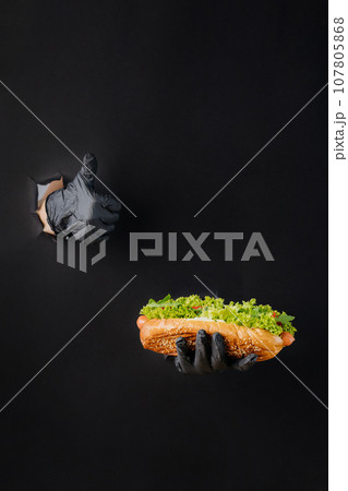Hand holding hot dog and showing a thumb up through torn holes in black paper background. Street food concept 107805868