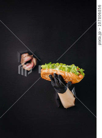 Female hand holding hot dog and looks through holes in black paper background. Street food concept Female hand holding hot dog and looks through holes in black paper background. Street food concept 107806856