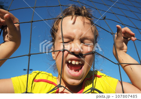 An 8-year-old Caucasian girl stands behind a sports net and screams. Volleyball sports dividing net against the blue sky close-up. Outdoor sports. 107807485