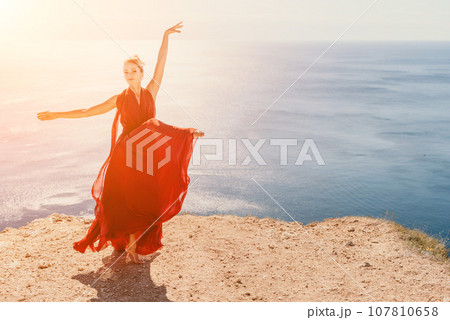 Woman in red dress on sea. Side view a Young beautiful sensual woman in a red long dress posing on a rock high above the sea on sunset. Girl on the nature on blue sky background. Fashion photo. Woman in red dress on sea. Side view a Young beautiful sensual woman in a red long dress posing on a rock high above the sea on sunset. Girl on the nature on blue sky background. Fashion photo. 107810658