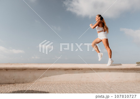 Fitness woman sea. Active, happy middle-aged woman in white sportswear exercises outdoors in a park over sea. Healthy lifestyle, physical activity and female fitness pilates and yoga routines. 107810727