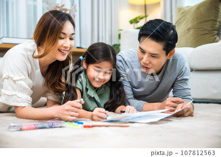 Smiling Father, Mother and daughter drawing together on paper at home, Asian family lying on floor painting with child daughter in living room, Happy family activity enjoy, learning draw art picture 107812563