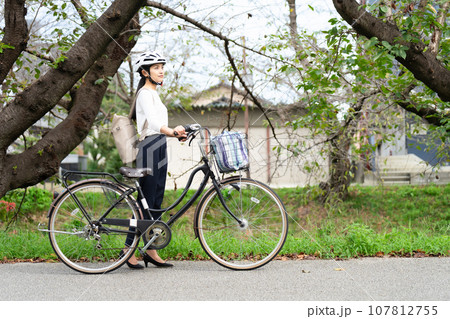 川沿いの遊歩道をサイクリングヘルメットをかぶって自転車を引いて歩く若い女性 107812755