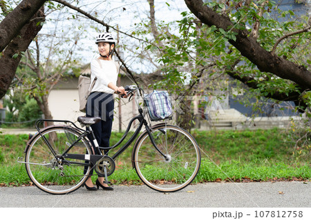 川沿いの遊歩道をサイクリングヘルメットをかぶって自転車を引いて歩く若い女性 107812758