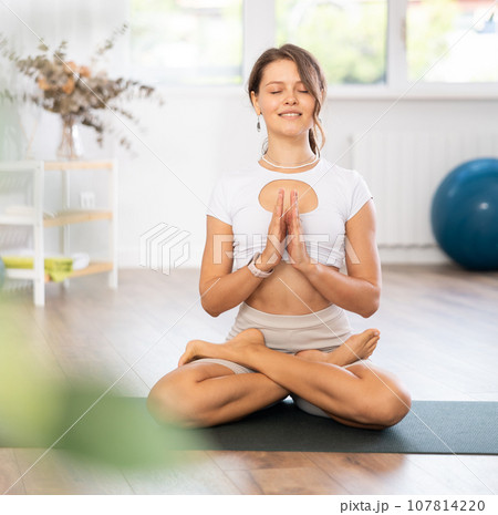 Young woman yoga trainer performing lotus pose variation on black rug Young woman yoga trainer performing lotus pose variation on black rug 107814220
