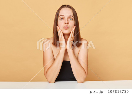 Romantic attractive Caucasian female model wearing black top sitting at table isolated over beige background posing with pout lips sending air kissing. Romantic attractive Caucasian female model wearing black top sitting at table isolated over beige background posing with pout lips sending air kissing. 107818759