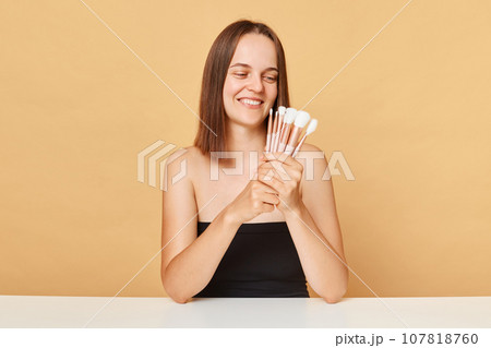 Beautiful brown haired woman with make-up brushes near attractive face smiling while looking away wearing black top posing with bare shoulders isolated over beige background. Beautiful brown haired woman with make-up brushes near attractive face smiling while looking away wearing black top posing with bare shoulders isolated over beige background. 107818760