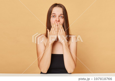 Shocked surprised Caucasian female model wearing black top sitting at table isolated over beige background looking at something astonishing covering mouth with palms. Shocked surprised Caucasian female model wearing black top sitting at table isolated over beige background looking at something astonishing covering mouth with palms. 107818766