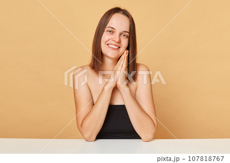 Cute charming joyful Caucasian female model with perfect skin wearing black top sitting at table isolated over beige background expressing positive emotions and good mood. 107818767